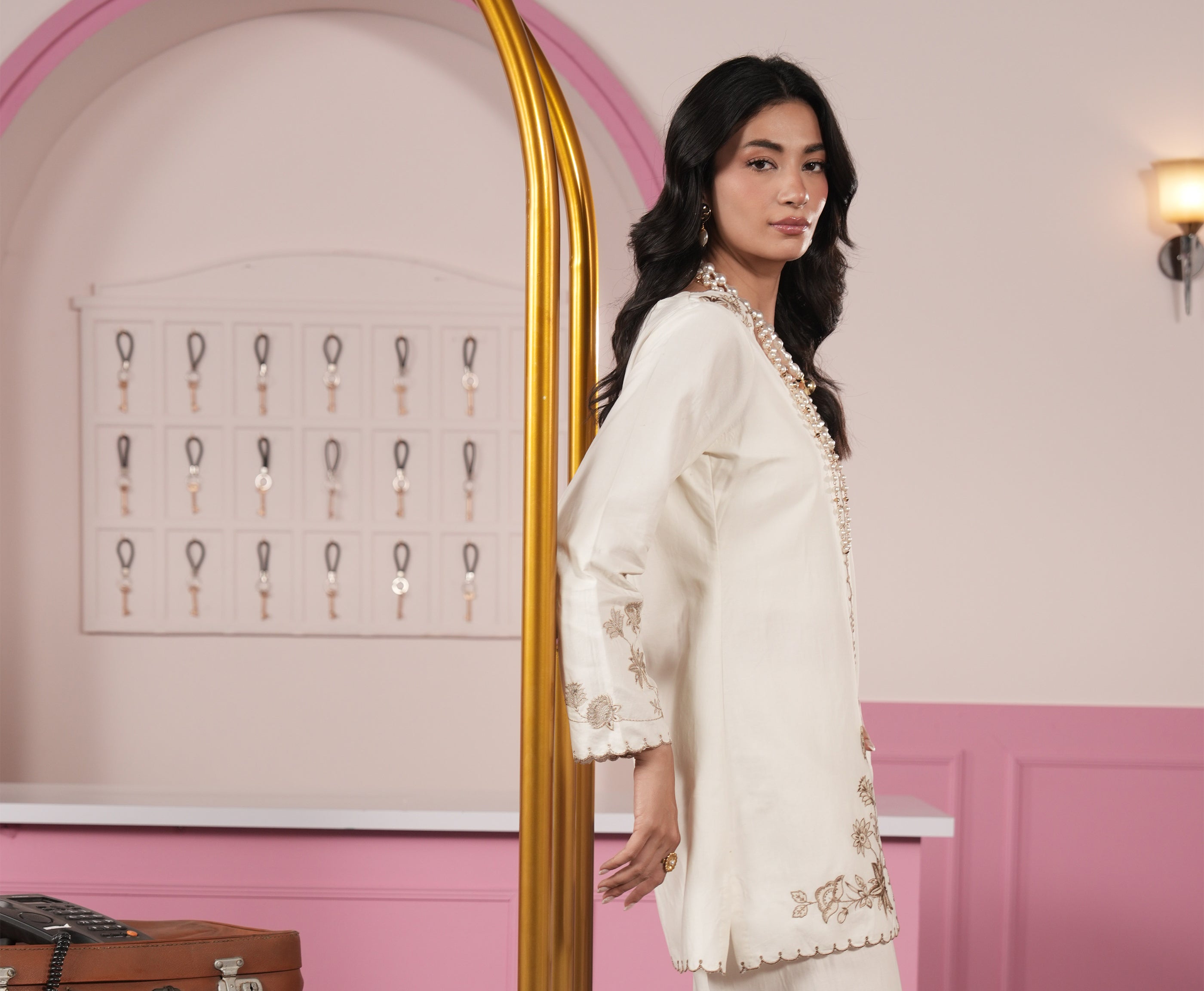 Woman in a white embroidered outfit standing in front of a pink wall with a mirror.
